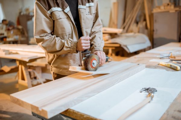 Handsome carpenter in uniform brushing wood with hand machine at the carpentry manufacturing